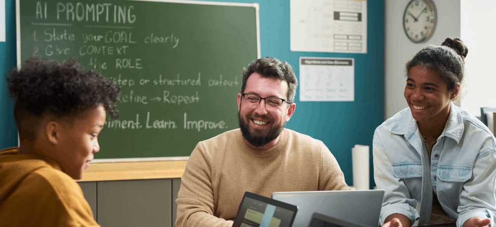 Teacher in classroom working with two students on computers. A chalkboard in the background shows guidelines for writing AI prompts.